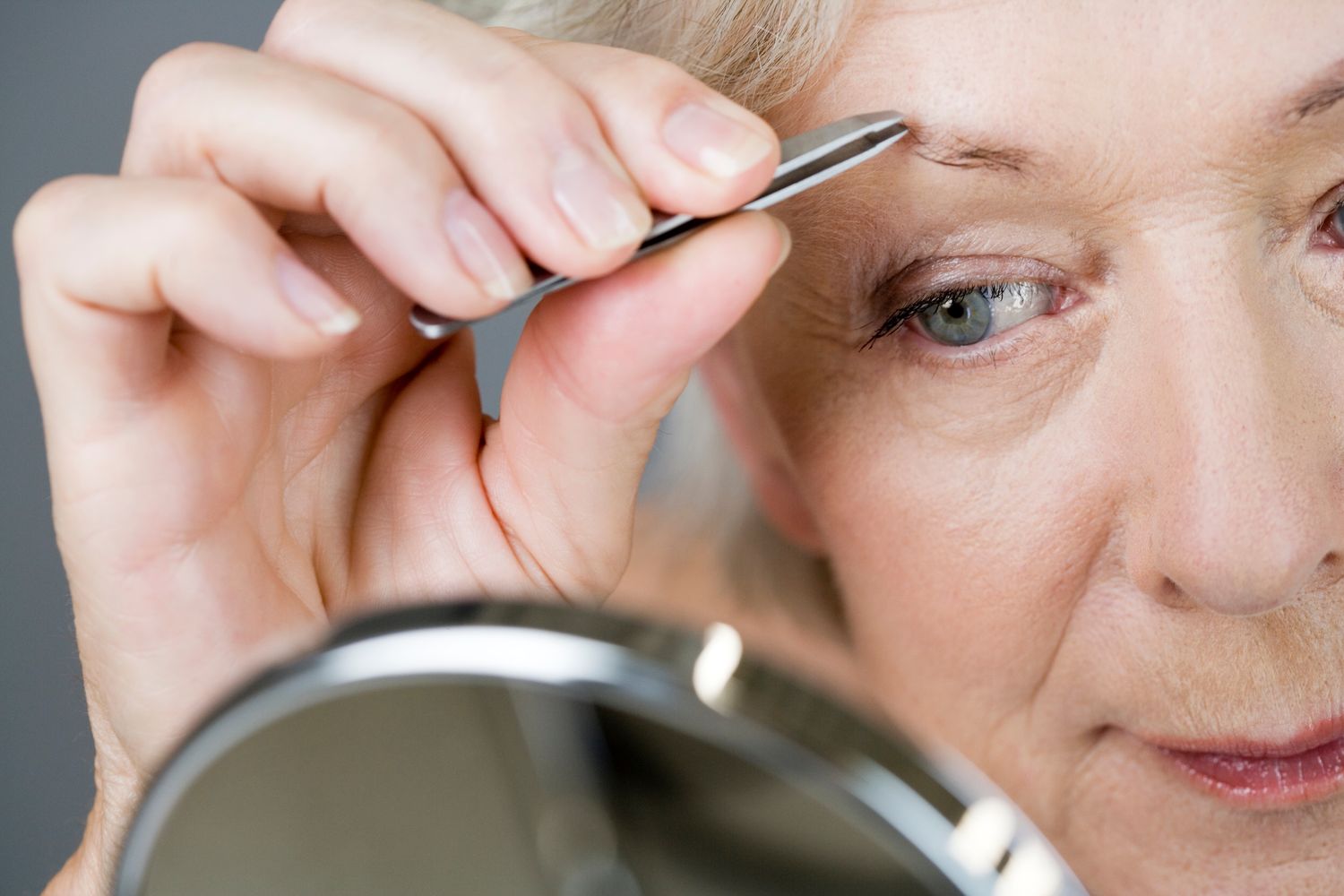 A mature woman tweezing her eyebrows with a hand mirror