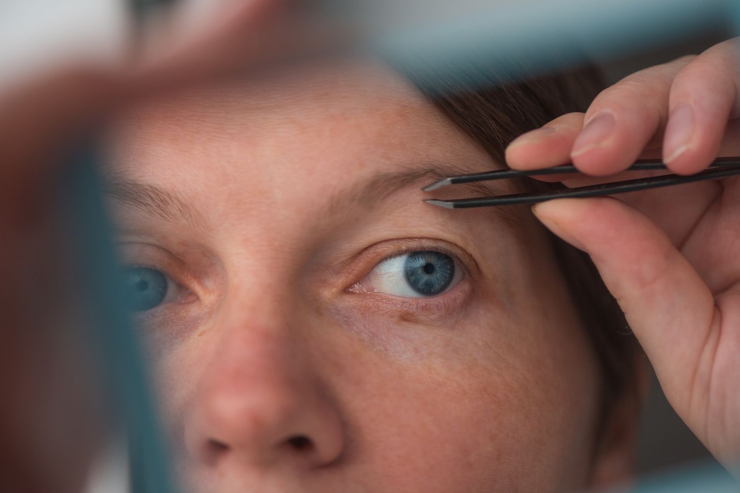 A woman using tweezers on her eyebrow in front of a mirror